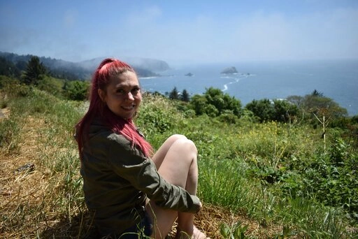 Alyssa outside sitting on the grass, overlooking a body of water while smiling